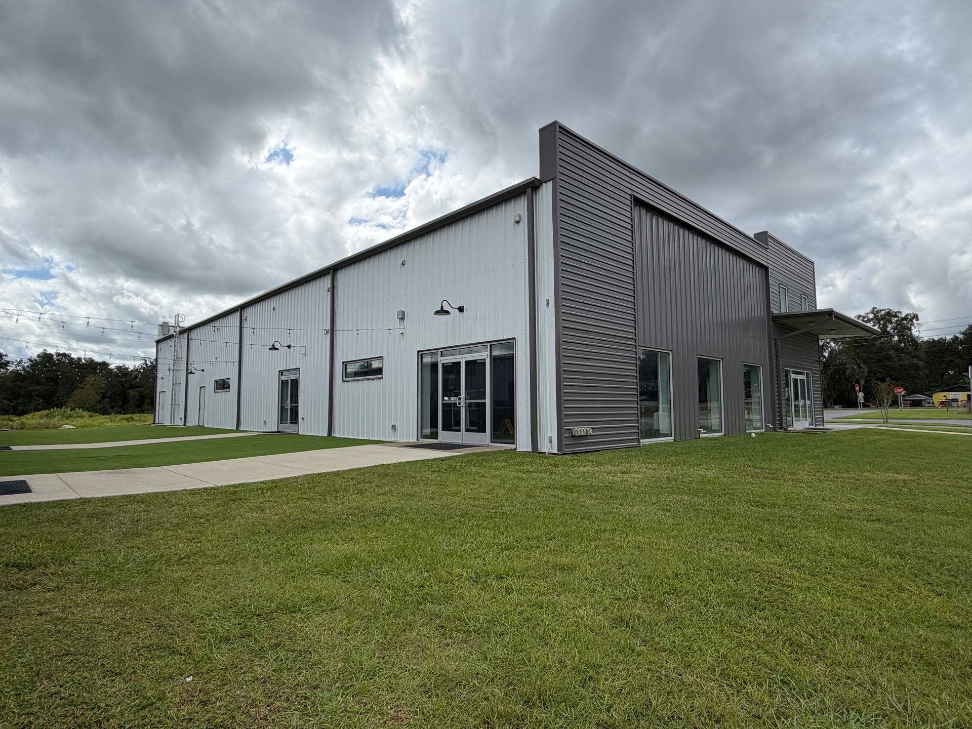 A modern, single-story building with a white metal side and a dark, corrugated facade, surrounded by green grass and trees.