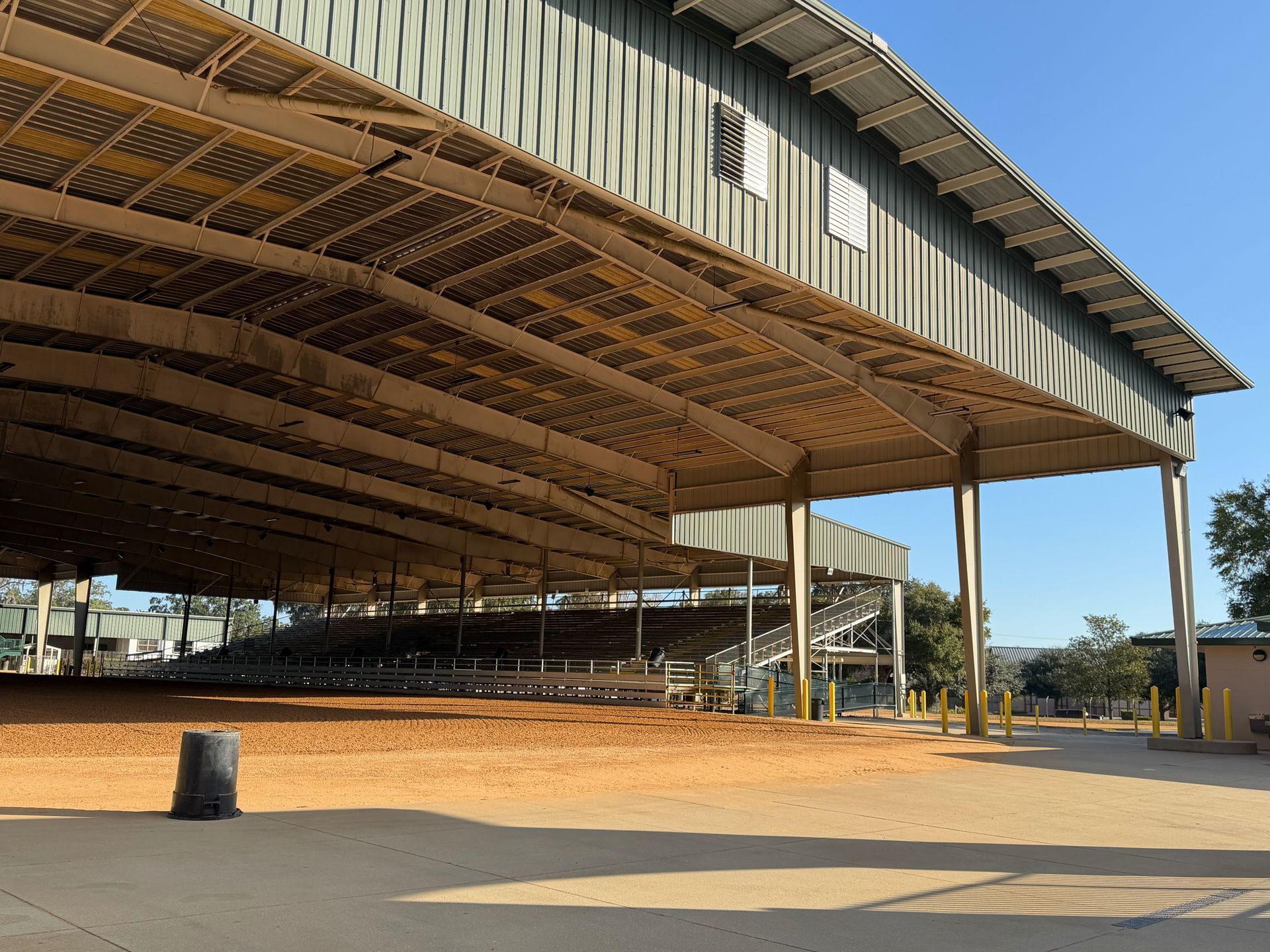 A large, open-air arena with a sand-covered ground and metal roofing, featuring tiered seating under the structure.