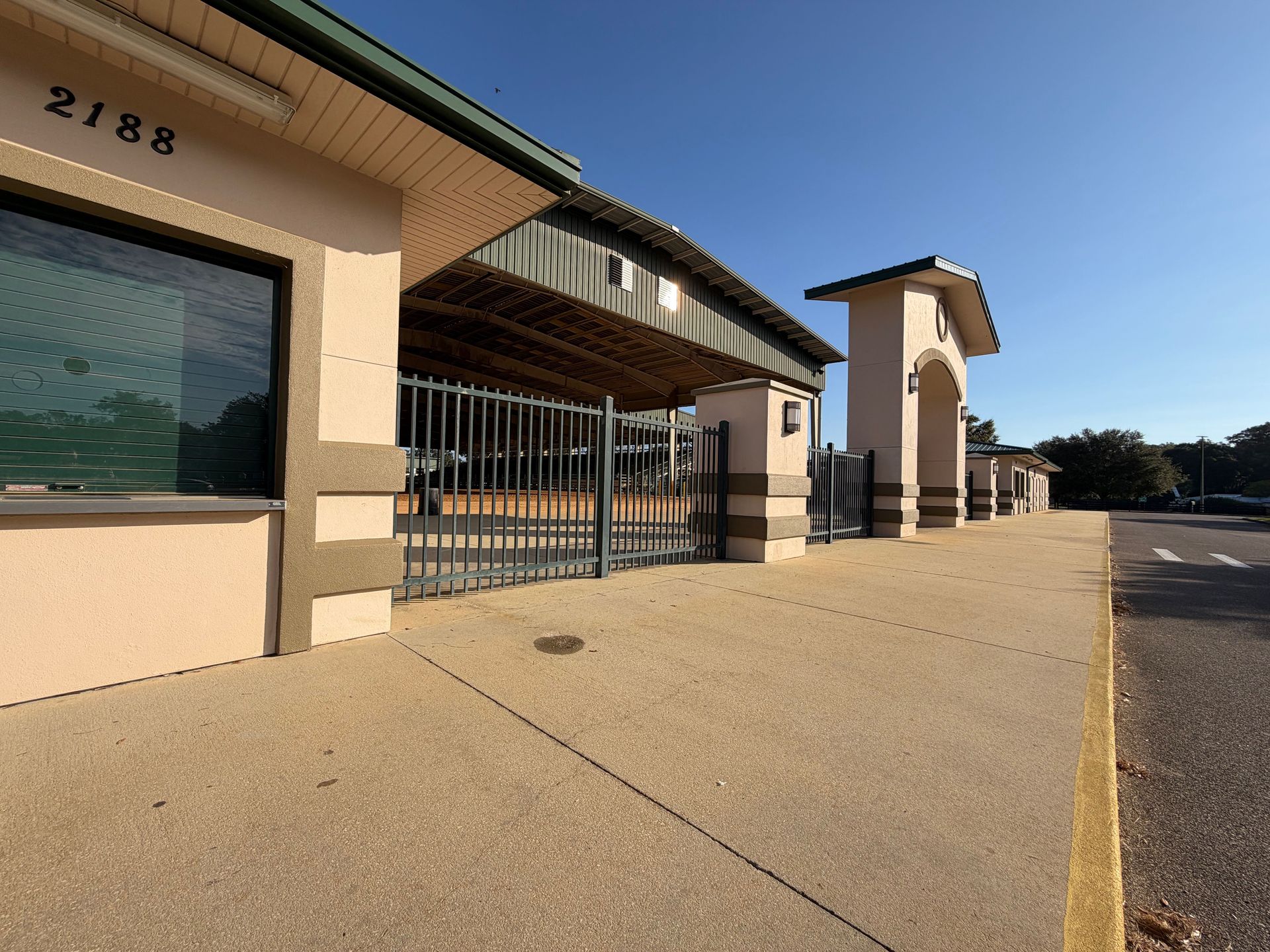 A sidewalk alongside a tan building with a gated, covered pavilion area under a clear blue sky.