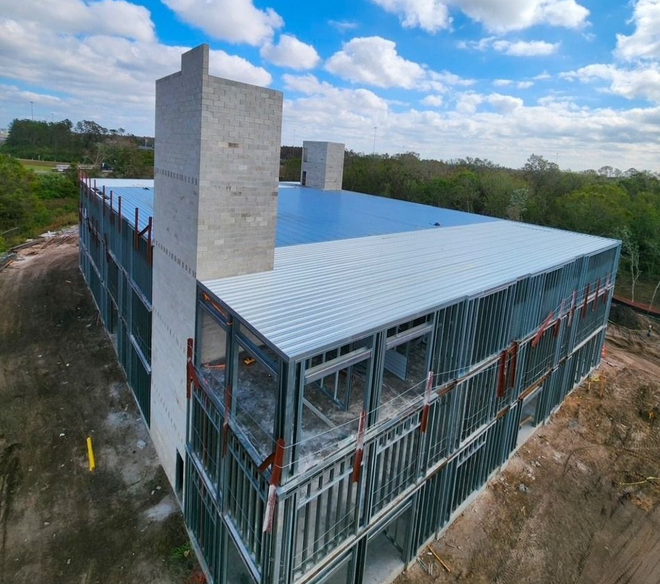 Aerial view of a steel-framed building under construction with a white roof and concrete towers, surrounded by trees