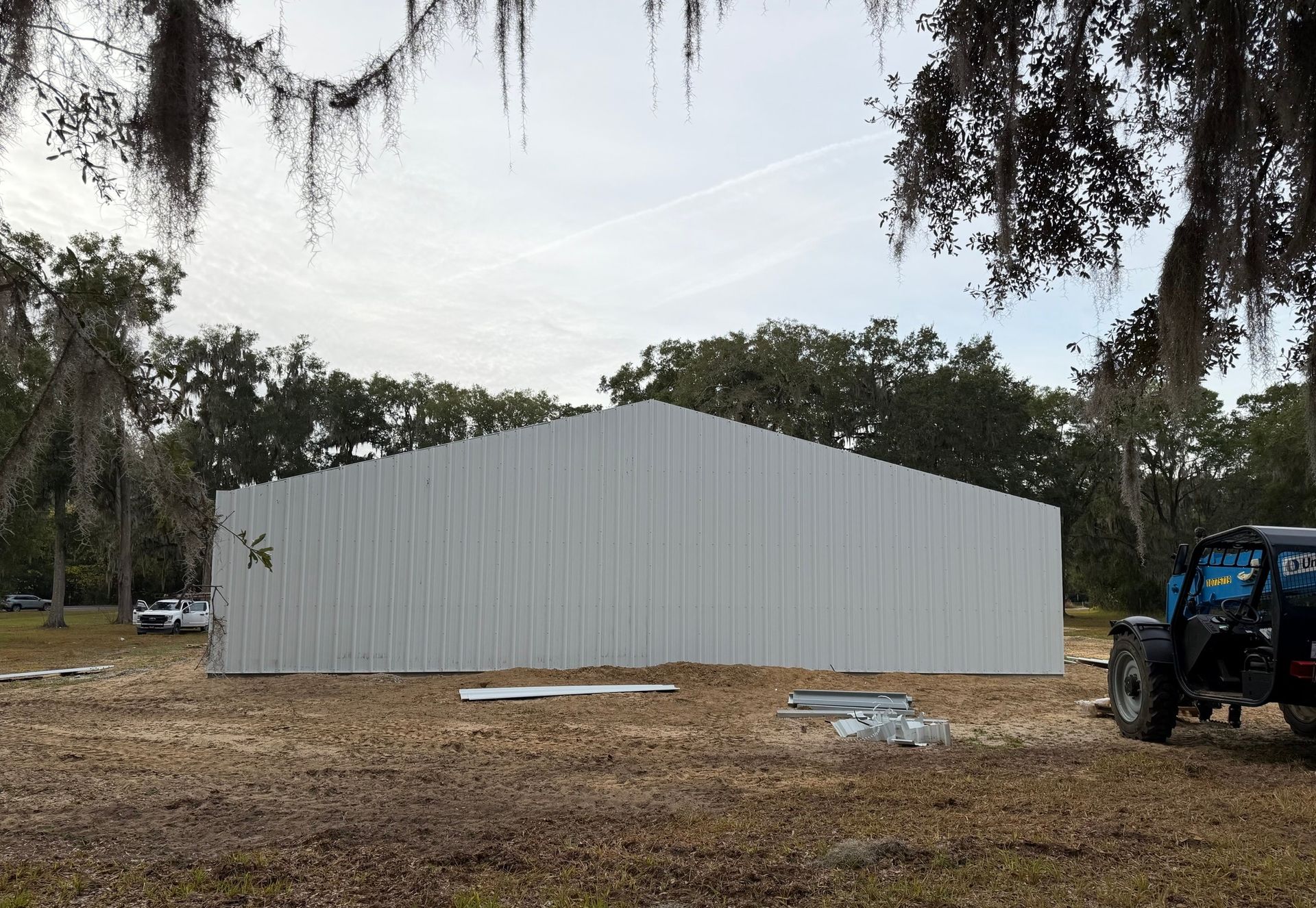 White metal building under construction on a wooded lot, with a blue tractor at right.