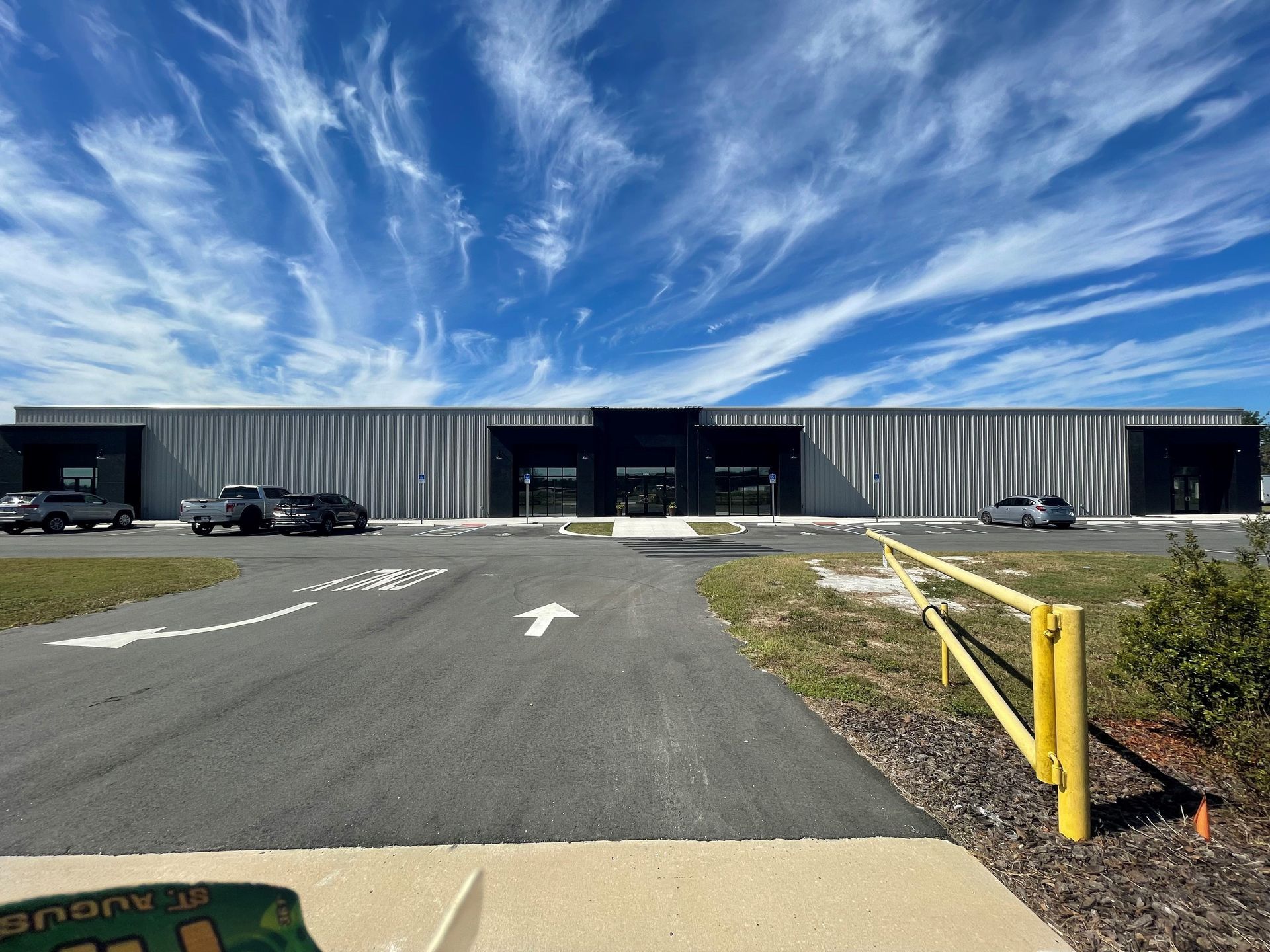 A modern, gray commercial building under a bright, cloudy sky with a paved parking lot and yellow guardrail in front.