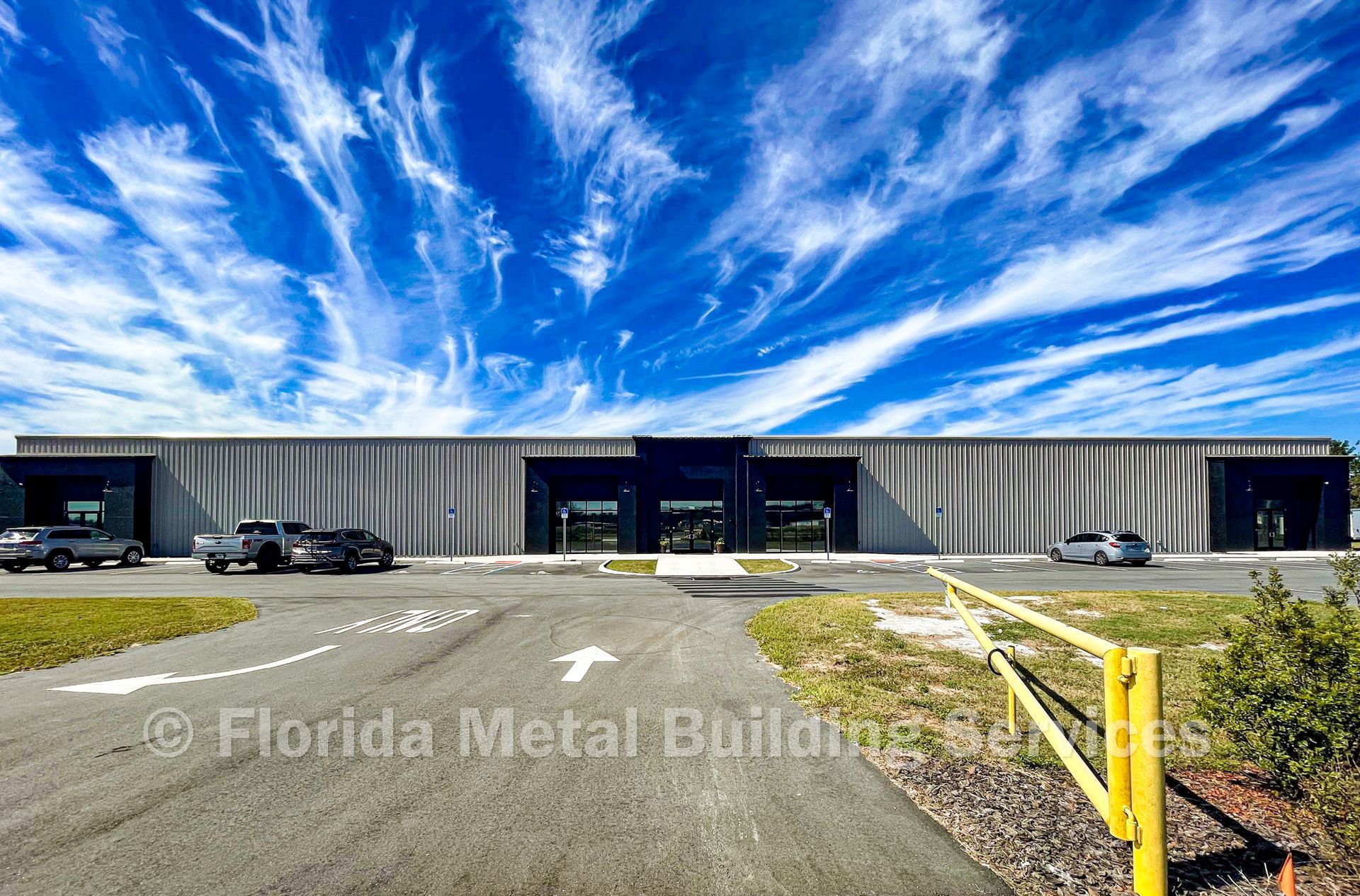 A wide, modern single-story commercial metal building under a bright, cloud-streaked blue sky.
