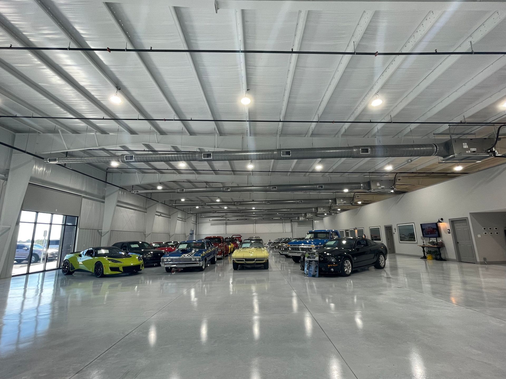A brightly lit warehouse display features a variety of classic and modern sports cars parked on a polished concrete floor.
