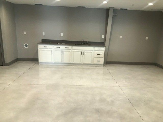 A white kitchenette cabinet with a dark countertop sits against a grey wall in a spacious room with polished concrete floors.