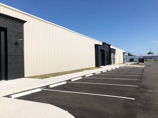 A long, beige metal-sided commercial building with black entryways and an adjacent paved parking lot under a blue sky.
