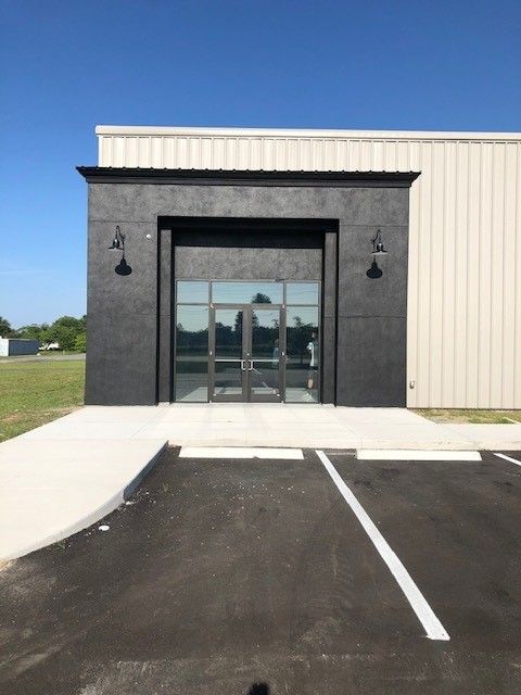 A modern building entrance with a prominent black-stucco facade, glass doors, and two outdoor lamps, set by a parking lot.