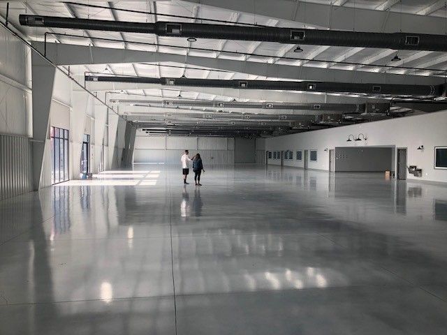Two people stand in a vast, empty, polished-concrete warehouse with high ceilings and white walls.
