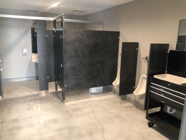 A public restroom featuring dark gray partition stalls, two wall-mounted urinals, a metal rolling cart, and concrete floors.