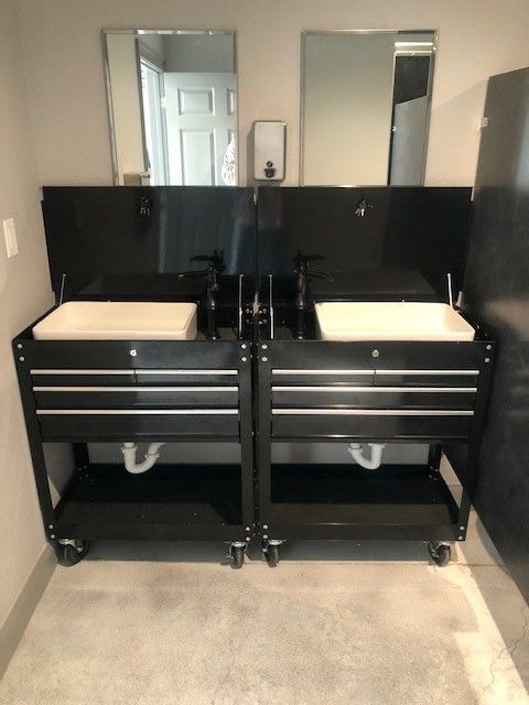 Two black tool chests converted into bathroom sinks, each with a white basin, faucet, and mirror, sitting on casters.