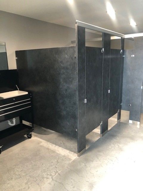 A row of dark grey bathroom stalls in a commercial restroom with concrete floors and a black utility cart nearby.