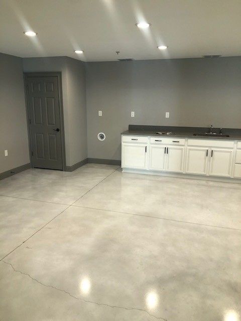 A finished basement with gray walls, a polished concrete floor, a dark gray door, and white cabinets with a countertop.