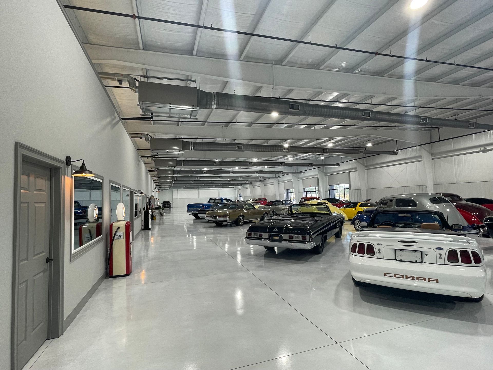 A bright, spacious indoor car showroom displaying several classic and modern vehicles on a polished concrete floor.