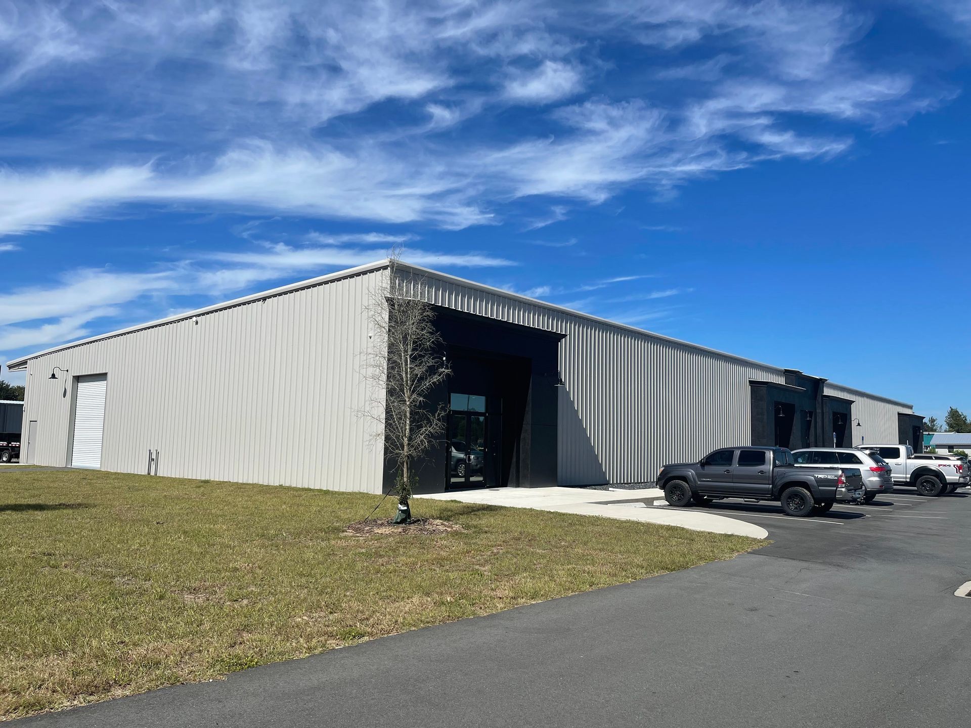 A wide, light-colored metal commercial warehouse building under a bright blue sky with parked pickup trucks in front.