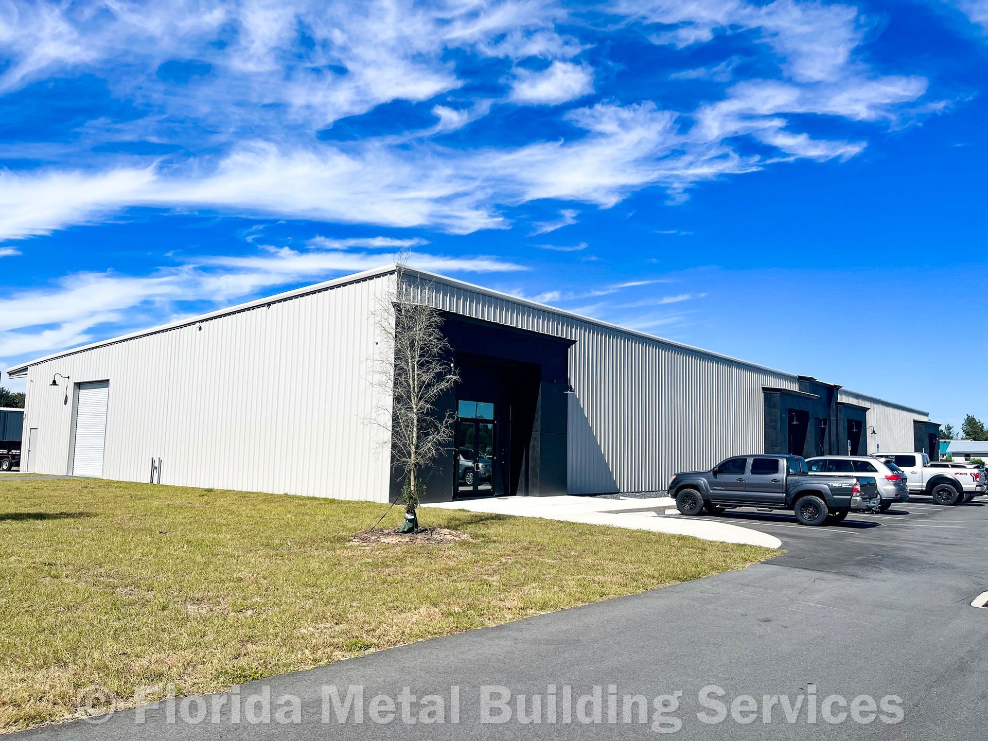 A long, one-story commercial metal building with a white exterior and dark entrance under a blue sky with scattered clouds.