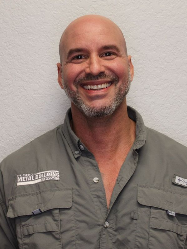 A smiling person with a shaved head and a gray beard, wearing a green button-down shirt with a branded chest patch.