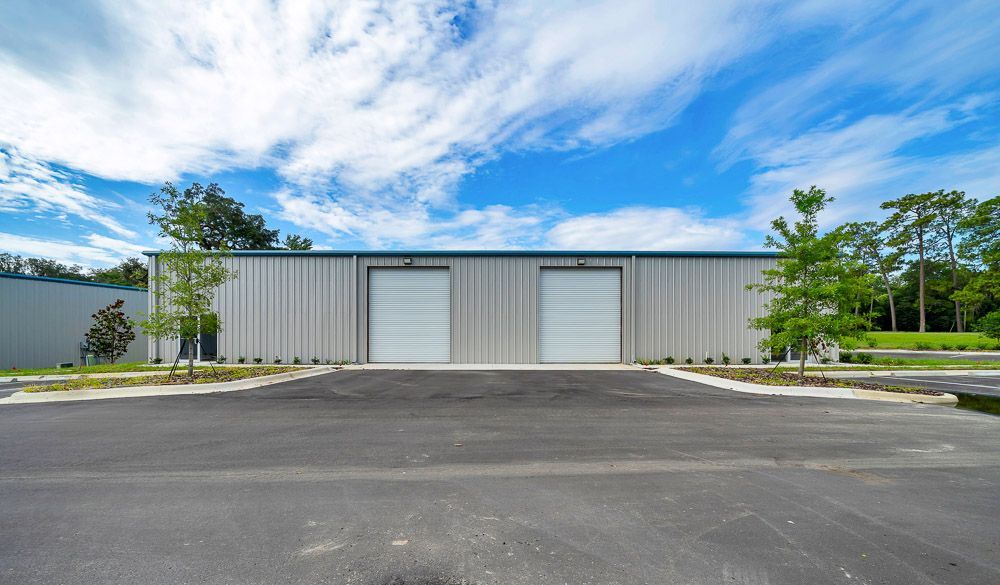 Gray industrial building with two garage doors, paved lot, and trees under a blue sky