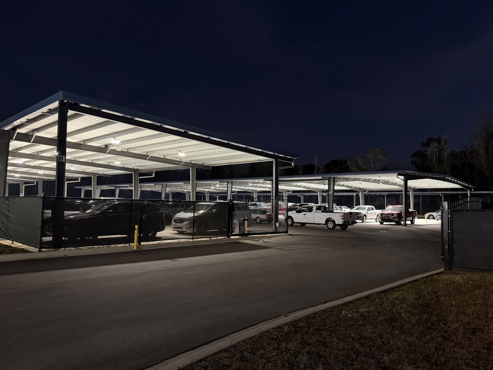 Rows of parked cars under illuminated metal solar panel carports in a paved lot at night.