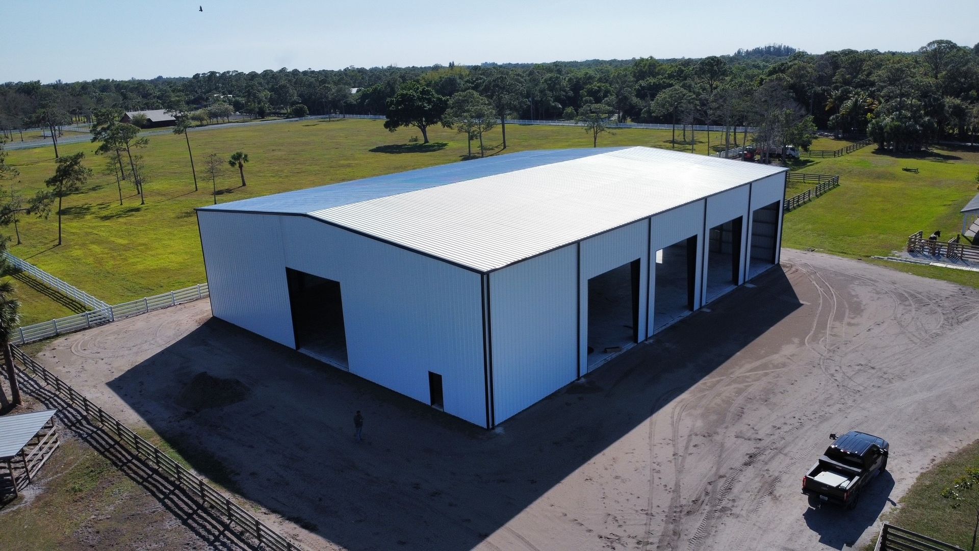 Aerial view of a white industrial warehouse on a paved lot in a rural area with a parked car nearby.