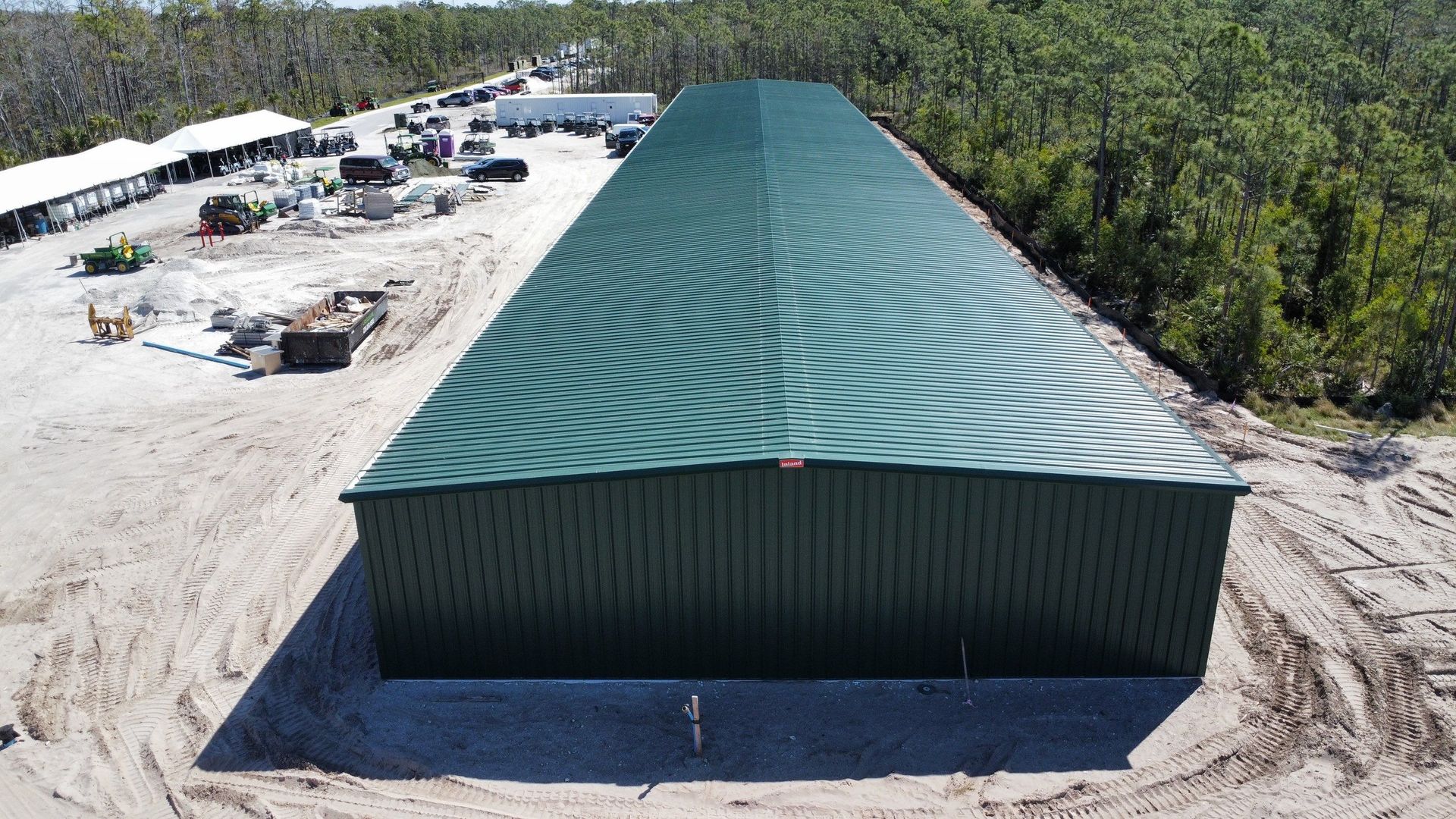 Aerial view of a large green-roofed warehouse beside a sandy lot and surrounding trees.