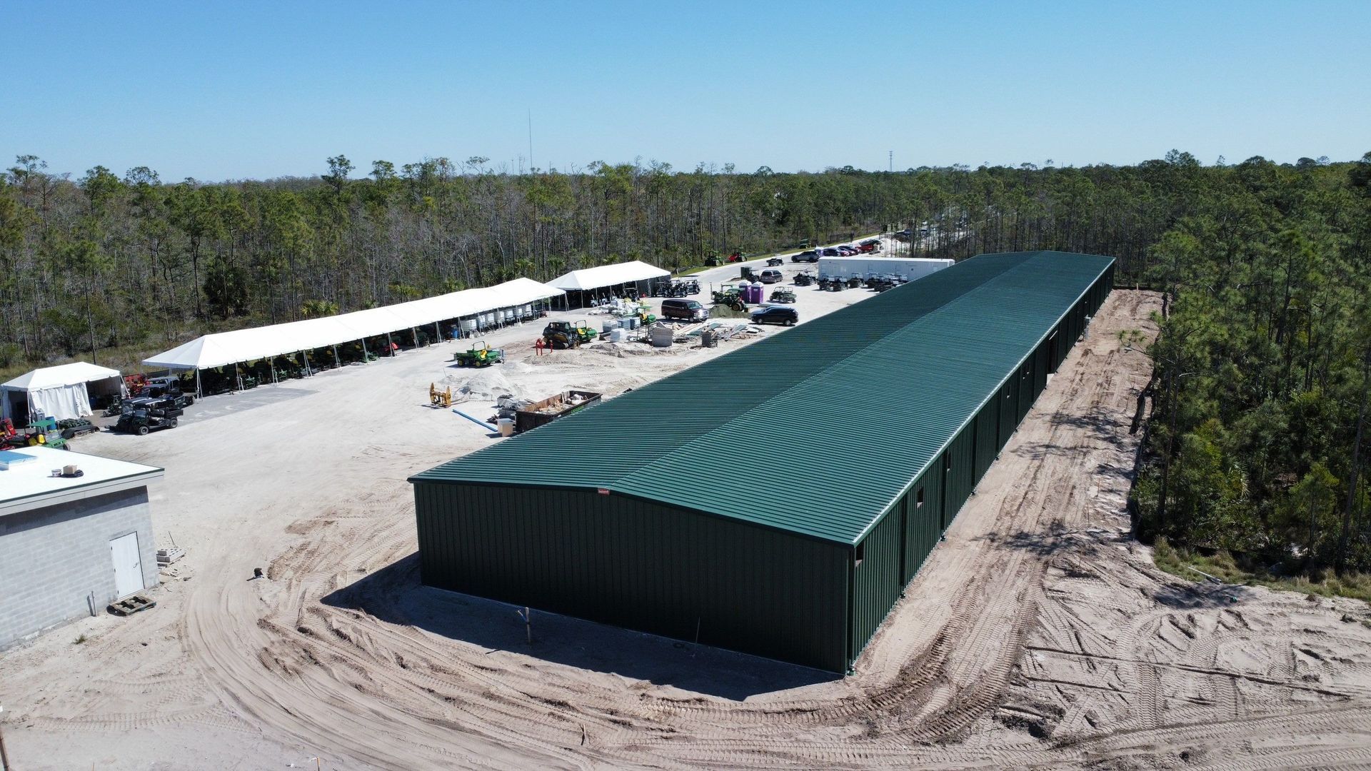 Aerial view of a long green-roofed warehouse under construction beside a sandy site and forest.