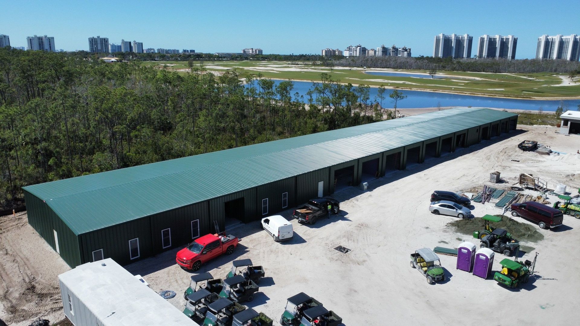 Long green-roofed building beside a gravel lot with parked vehicles and equipment near wetlands and distant towers.
