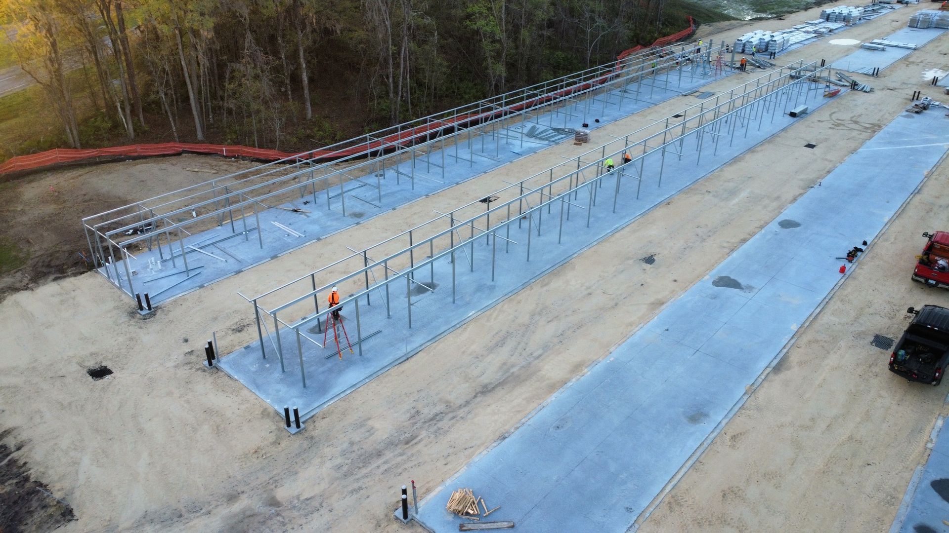 An aerial view of a construction site showing two long concrete foundation pads with metal frame structures being erected.