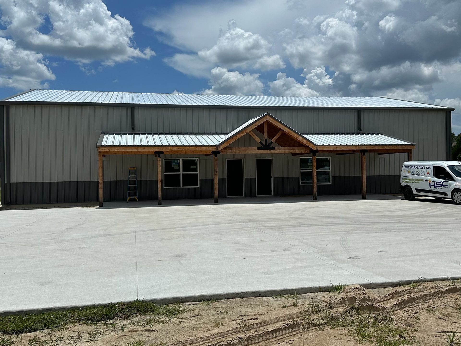 Large gray metal building with a covered front entrance, concrete lot, and a white van parked at right under a cloudy sky.