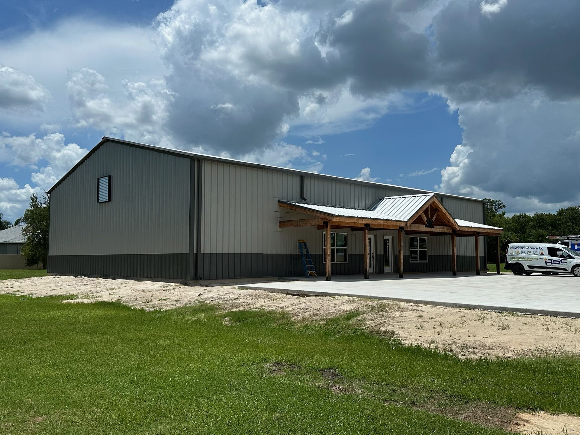 Large gray metal building with a covered entrance, parked van, and cloudy sky over a grassy lot.