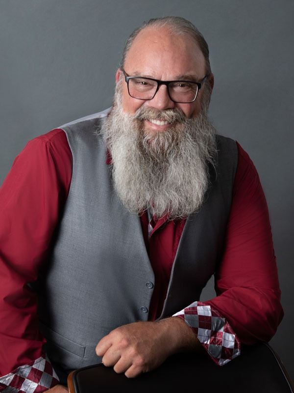 A smiling person with a long, gray beard, wearing a red shirt, gray vest, and checkered cuffs, against a gray background.