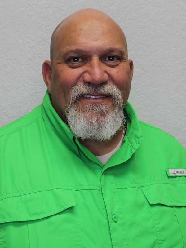 A person with a grey goatee wearing a bright green, short-sleeved button-down shirt, smiling against a plain white wall.