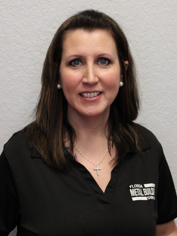 A smiling person with shoulder-length brown hair, wearing a black collared shirt with a logo and a silver cross necklace.