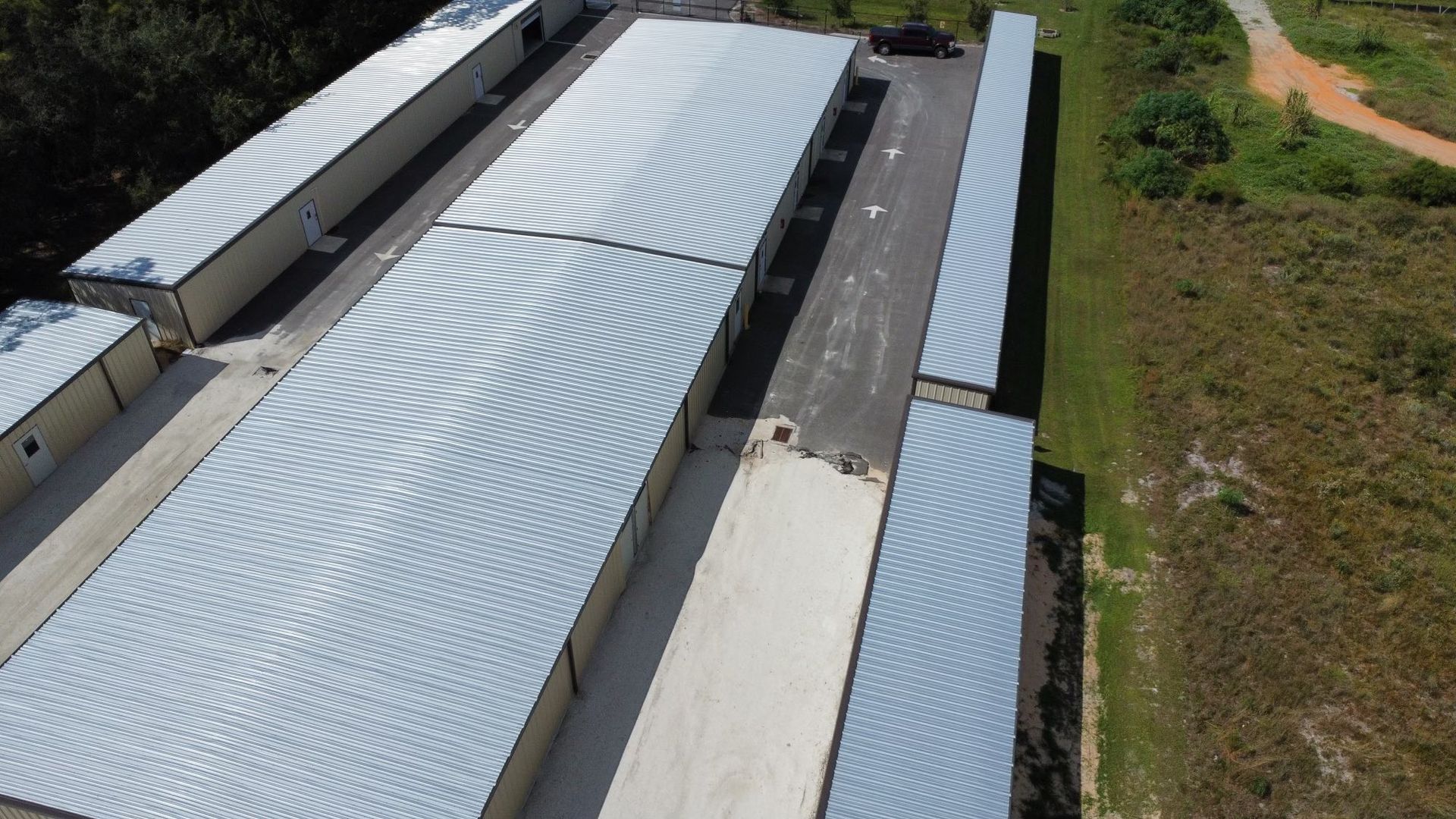 Aerial view of long metal-roofed industrial buildings beside a dirt road and green field.