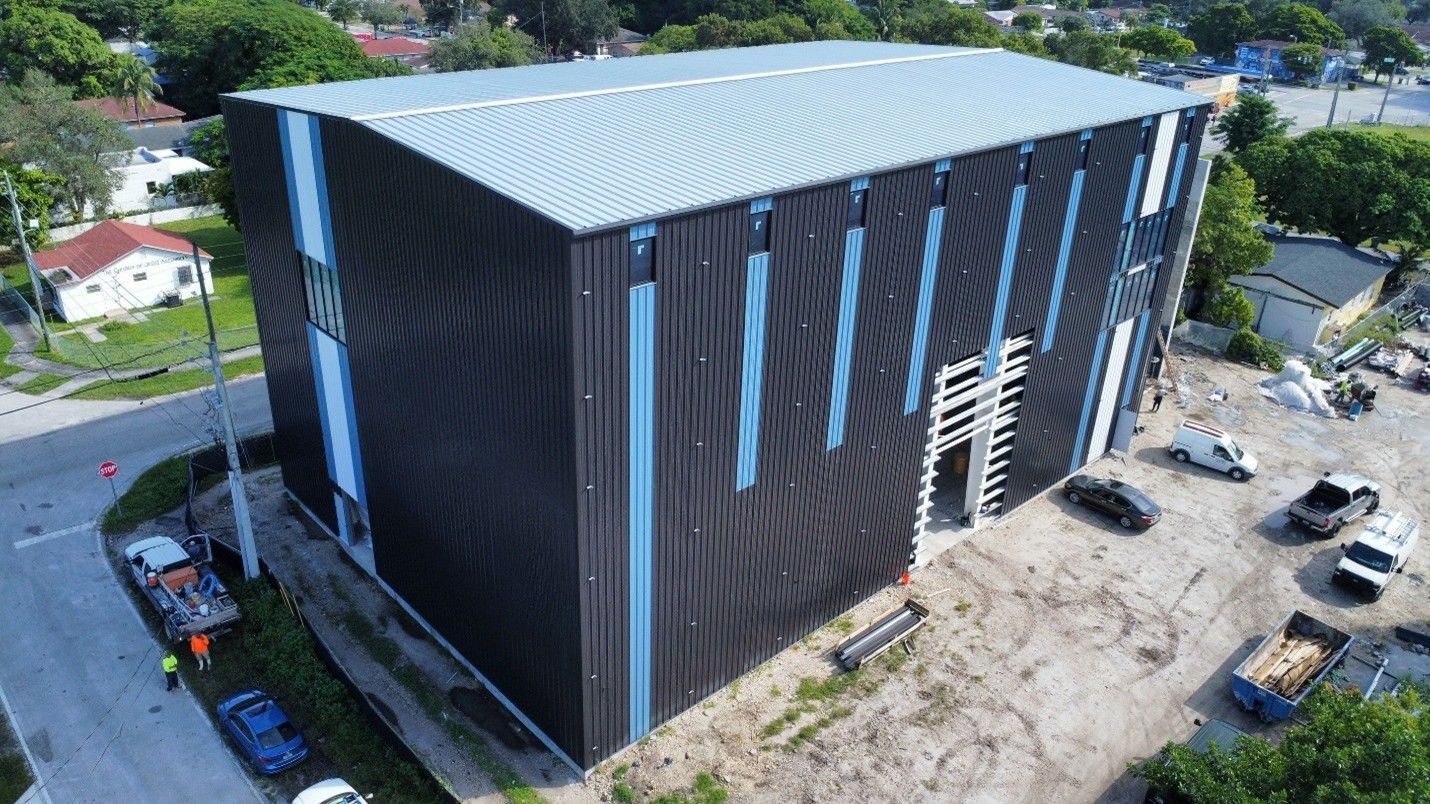 Aerial view of a large black building with blue vertical accents and a gray roof at a construction site.