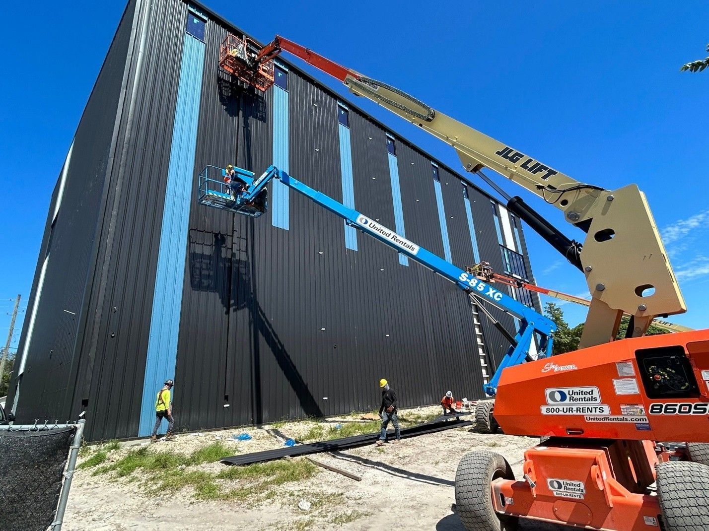 Workers and cranes install panels on a tall black building exterior under a clear blue sky.