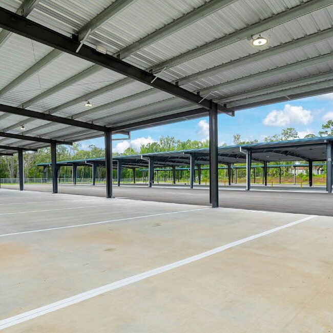 An empty, modern parking lot with concrete stalls sheltered by a metal roof structure under a bright, partly cloudy sky.