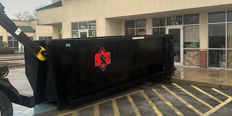 A black dumpster is being unloaded in front of a building. The building has large windows and is made of beige bricks.
