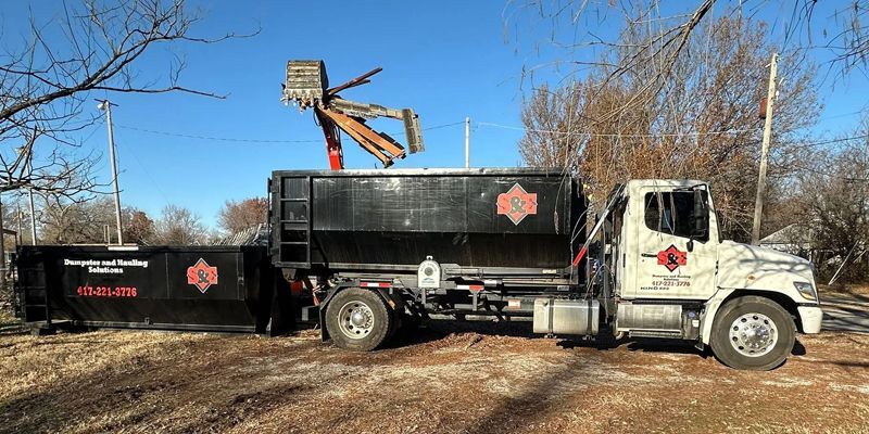 A black dumpster truck with a raised arm, used for debris removal, on a sunny day.