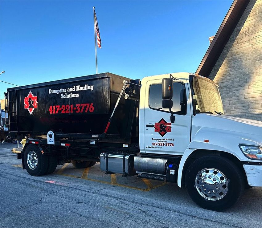 White and black dumpster truck parked on pavement with building in background; phone number visible.