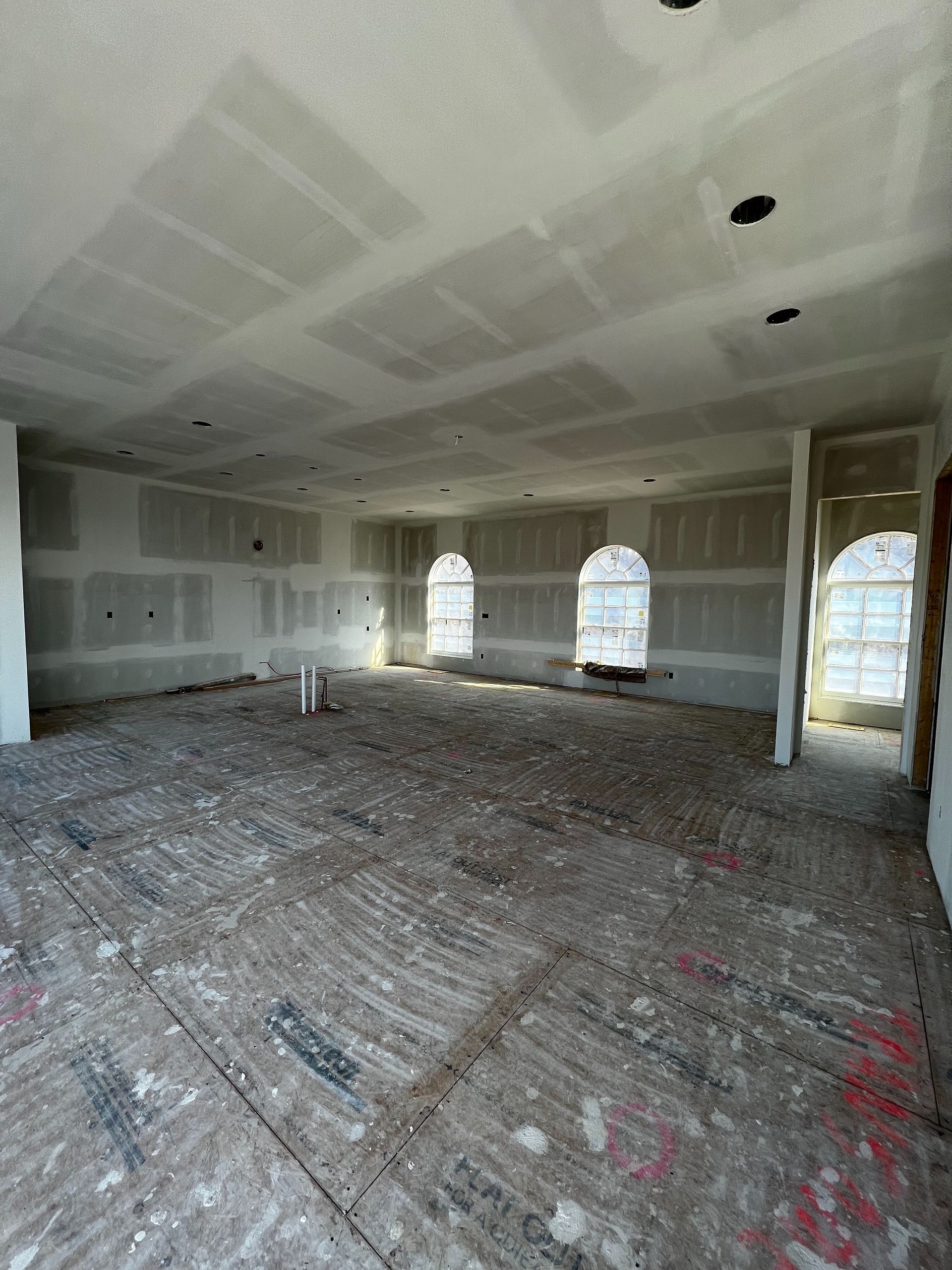 Interior of a room under construction with exposed floorboards, drywall, and arched windows.
