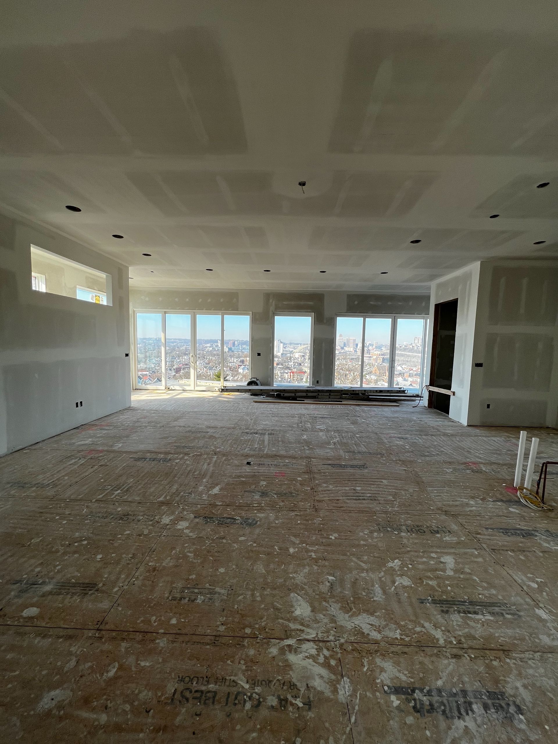 Interior view of a large room under construction; drywall, windows with city view, unfinished floor.
