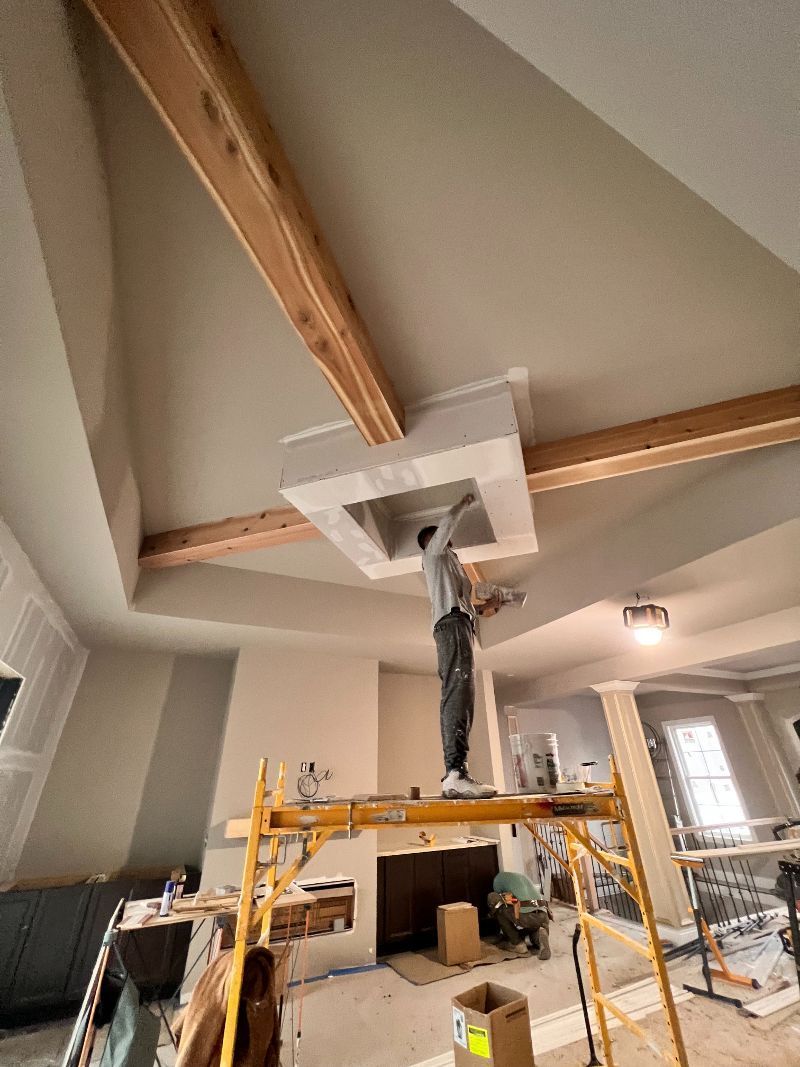 Man on a scaffold working on a white architectural feature on a high, light-colored ceiling with wooden beams.