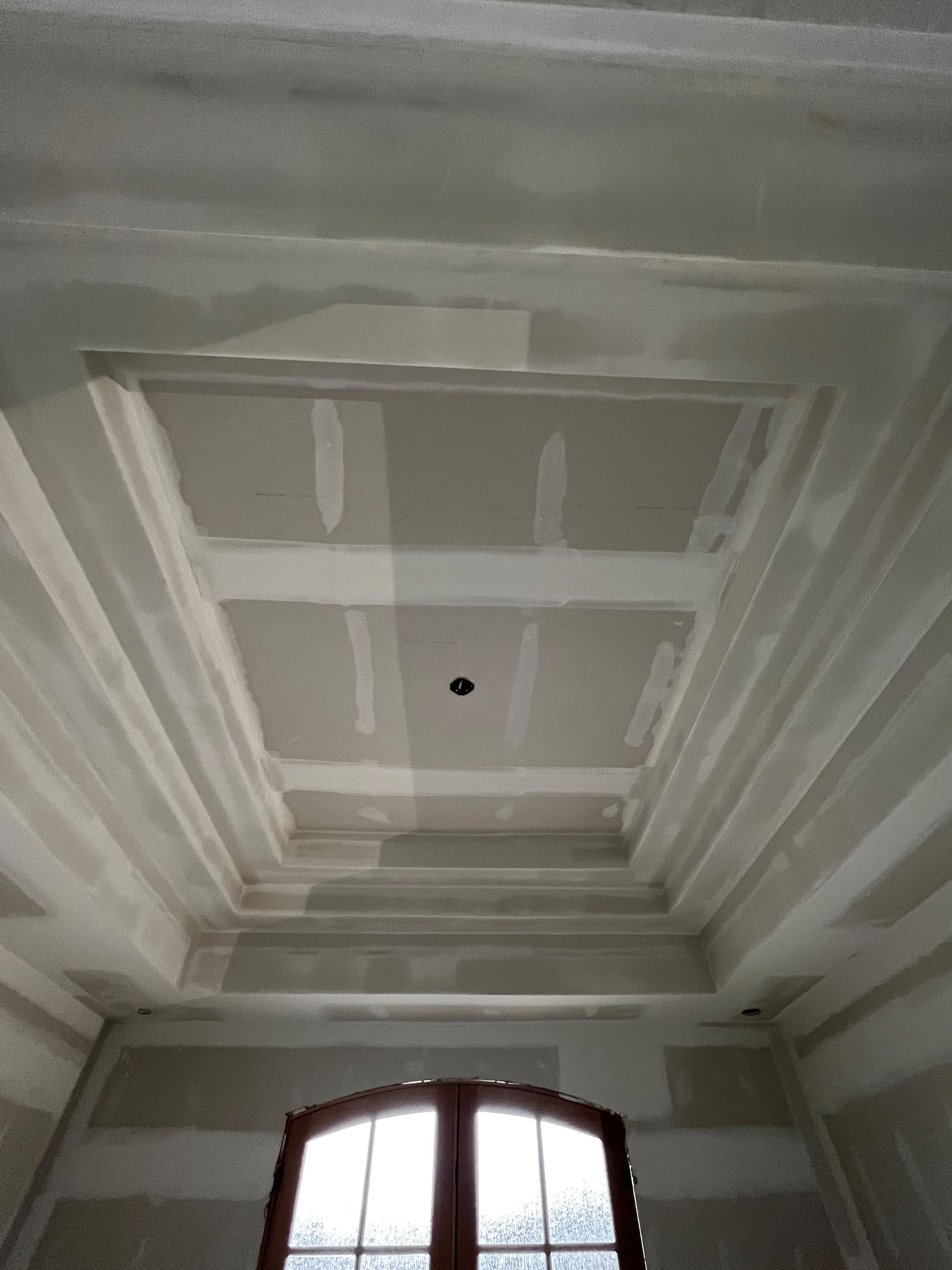 Drywall ceiling with recessed panels and a central light fixture above a window.