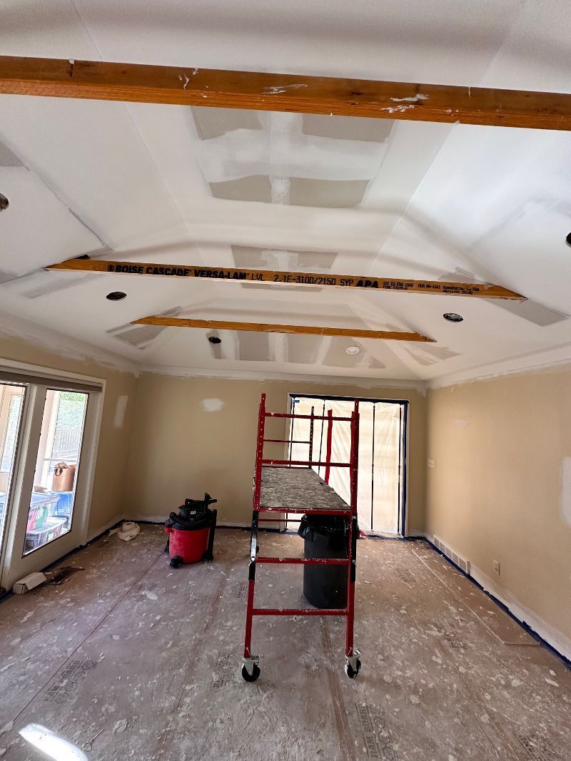 Room under renovation with scaffolding, exposed beams, and drywall. Red vacuum cleaner and black trash can present.