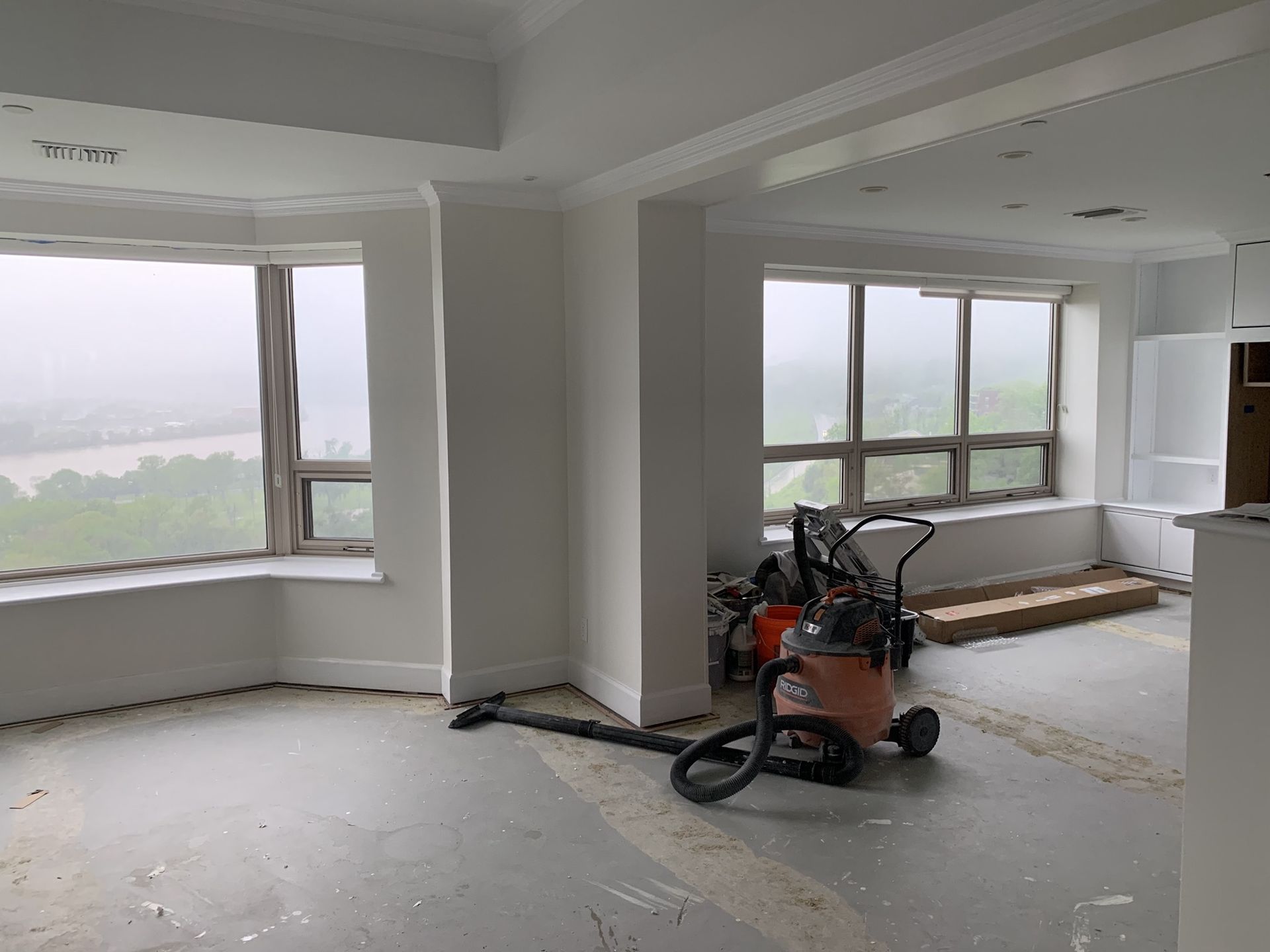 A mostly white, unfinished room with large windows overlooking a foggy cityscape. Vacuum cleaner sits in the center.