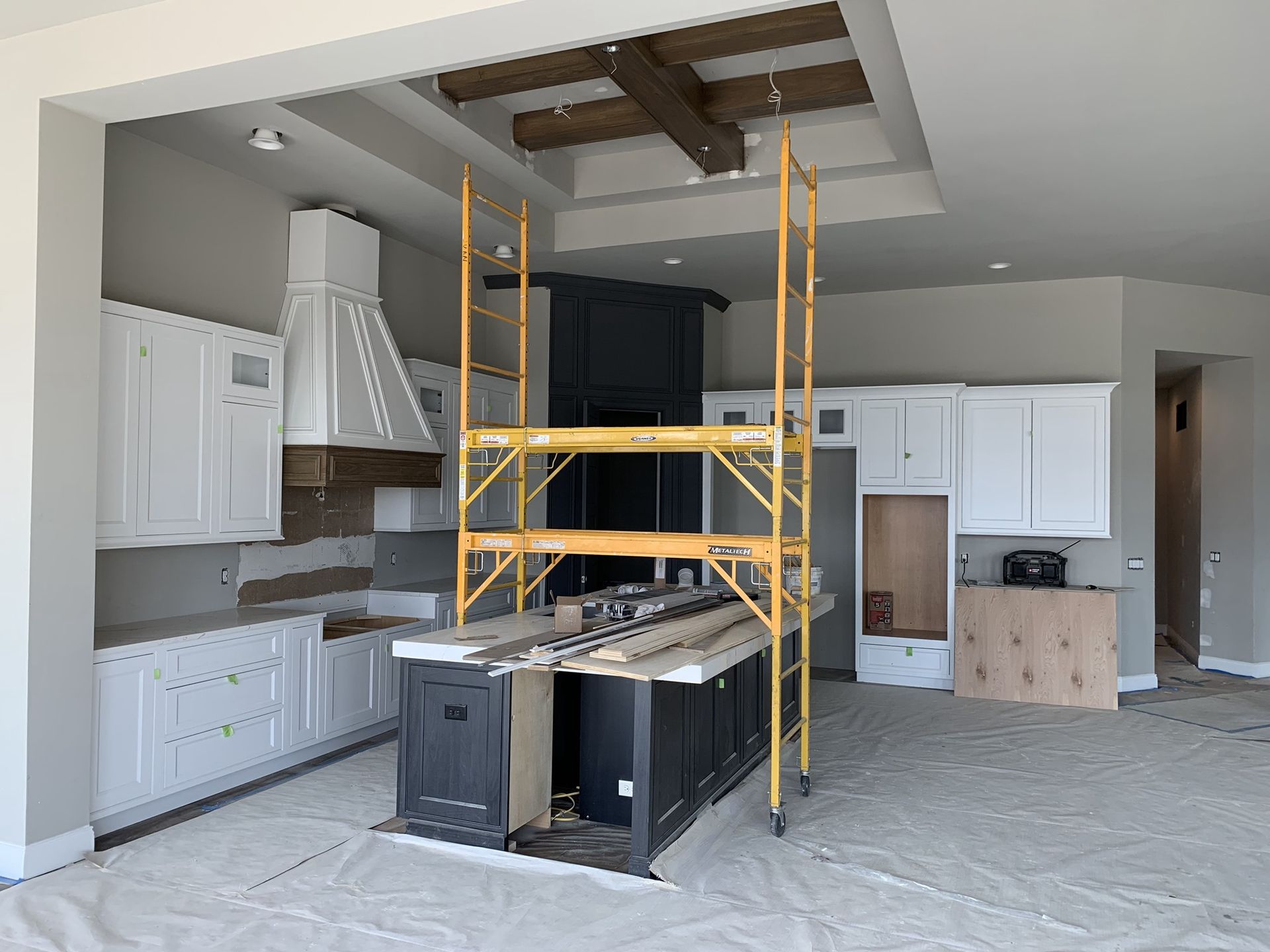Kitchen remodel: White cabinets, black island, wooden ceiling beams, scaffolding, gray walls, and light floor.