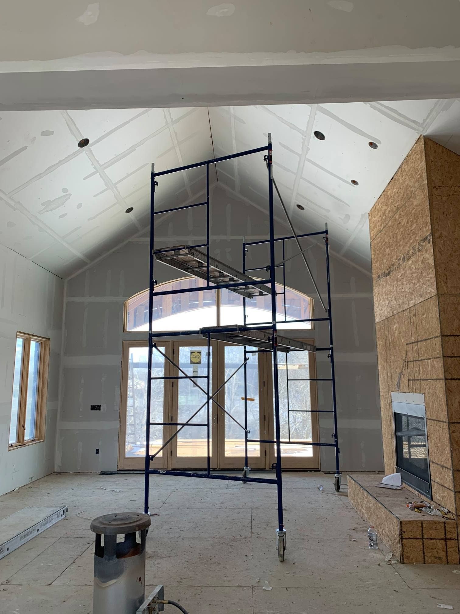 Interior of a house under construction, drywall, scaffolding, large windows, and a fireplace.