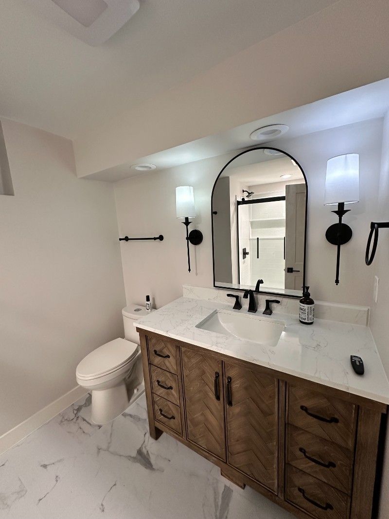 Bathroom with wood vanity, marble countertop, arched mirror, black fixtures, and white walls.