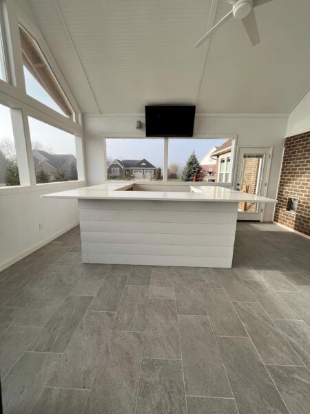 White sunroom with a large island, TV, and gray tile floor. Large windows provide views of houses.