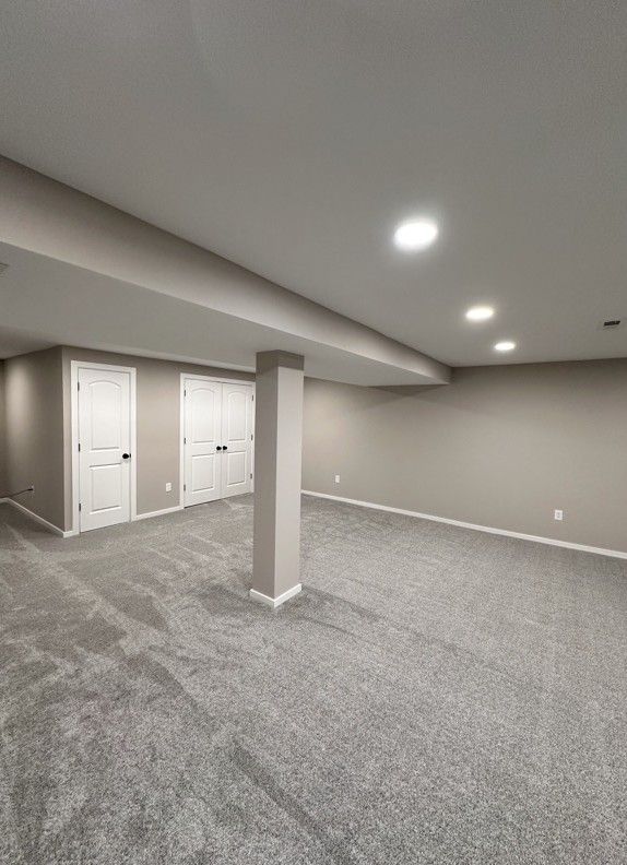 Empty, carpeted basement with white doors and a support column. Walls are painted beige, with recessed lighting.